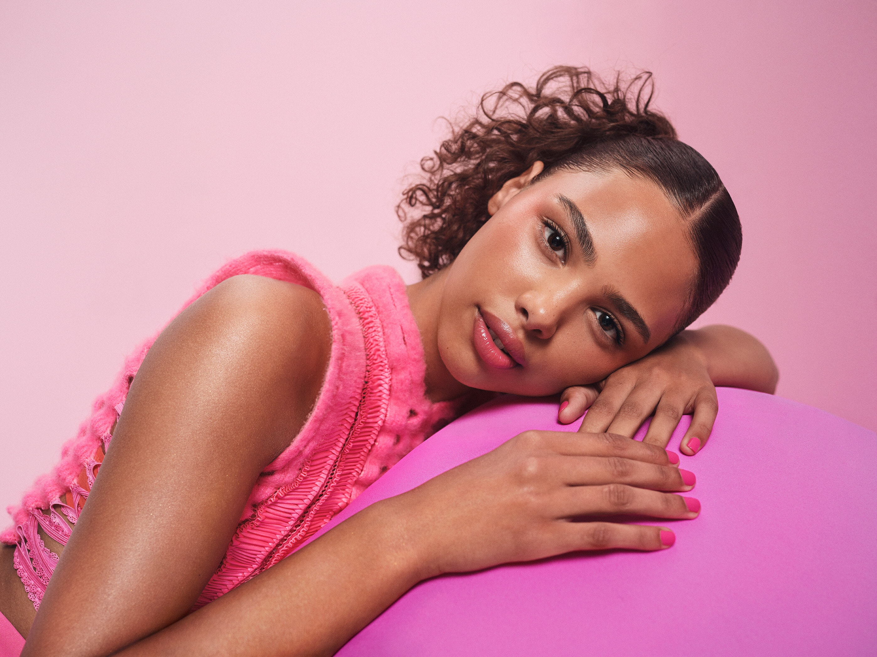 一名女性臉部靠在粉紅色圓形物品上，眼神柔和，臉上帶著粉紅色的 MAC 超漾光幻色嫩唇凍及超漾光持色奶凍腮紅，整體妝容顯得自然迷人。 